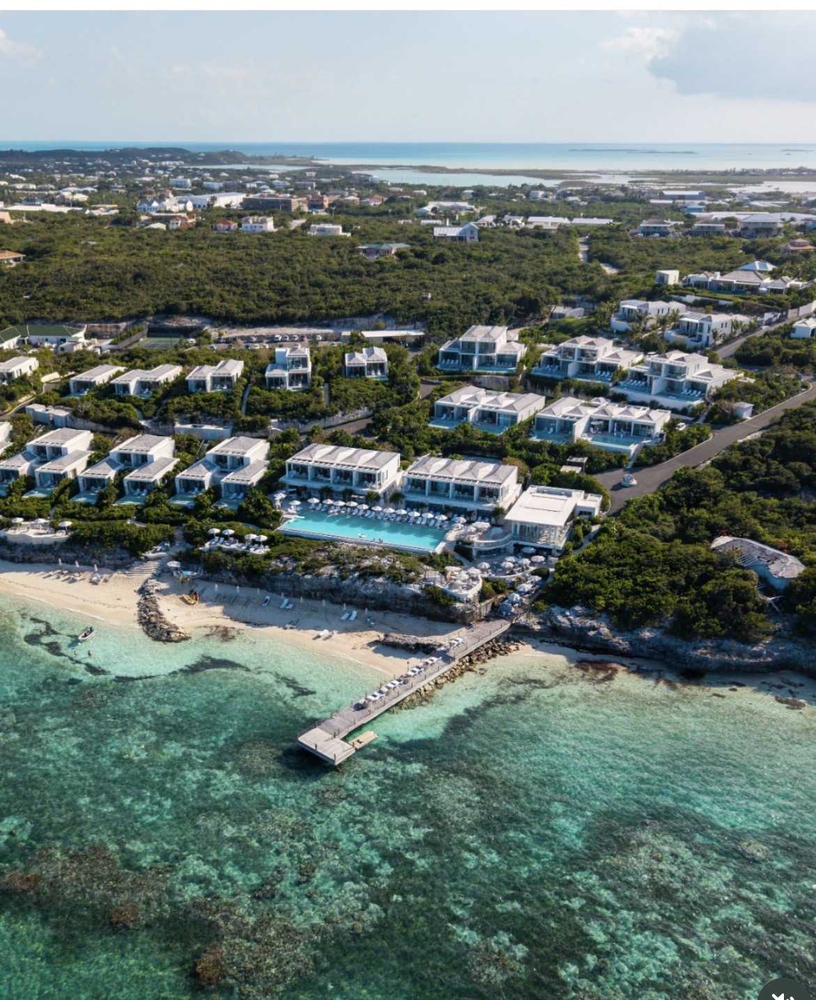 Aerial view of a white beachfront resort with villas, pool, and dock over turquoise reef water